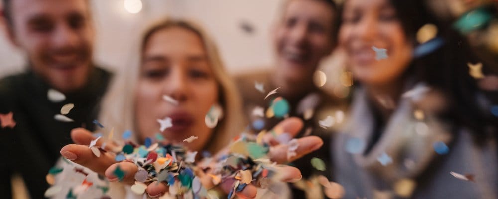 A blonde woman blows colorful confetti from her hands during a festive celebration with friends.