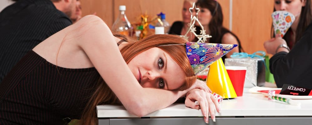 A red-haired woman with a party hat rests her head on a table, looking bored at an office party.